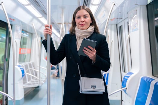 Stylish young woman in a black coat and beige scarf reading on an e-reader while standing inside a modern subway train. She balances while holding onto a pole during her commute.