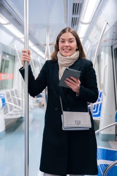Stylish young woman in a black coat and beige scarf reading on an e-reader while standing inside a modern subway train. She balances while holding onto a pole during her commute looking at camera