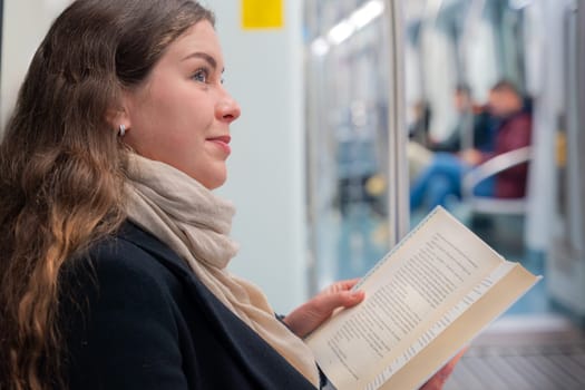 Young woman in a black coat and beige scarf sitting inside a modern subway train, reading a book with focus. She enjoys a quiet moment during her commute in the city.