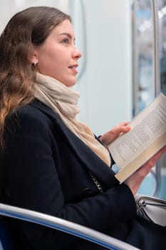 Young woman in a black coat and beige scarf sitting inside a modern subway train, reading a book with focus. She enjoys a quiet moment during her commute in the city.