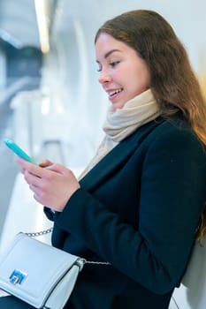 Stylish young woman in a black coat and scarf sitting in a modern subway station, smiling while using her smartphone. The bright and clean environment enhances the urban setting.