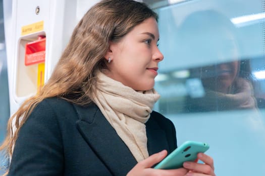 Thoughtful young woman in a black coat and scarf holding her smartphone while gazing out the window of a subway train. The urban commute setting reflects contemplation and modern connectivity.