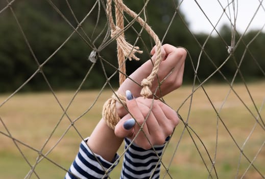 Two hands hold onto a knot in a net, with green grass and trees in the background. The fingers display nail polish, indicating a casual moment in nature.