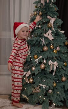 Christmas concept. a 5-year-old boy in New Year's pajamas and a Santa Claus hat stands next to the Christmas tree and joyfully decorates it. Happy smile.