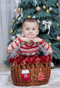 Christmas. A five-year-old boy in New Year's pajamas sits on the floor near a wicker New Year's basket against the backdrop of a Christmas tree and smiles cheerfully and makes faces. Facial expressions festive atmosphere