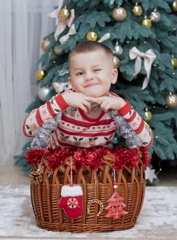 Christmas. A five-year-old boy in New Year's pajamas sits on the floor near a wicker New Year's basket against the backdrop of a Christmas tree and smiles cheerfully and makes faces. Facial expressions festive atmosphere
