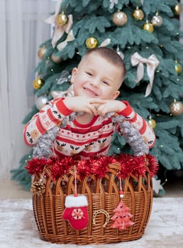 Christmas. A five-year-old boy in New Year's pajamas sits on the floor near a wicker New Year's basket against the backdrop of a Christmas tree and smiles cheerfully and makes faces. Facial expressions festive atmosphere