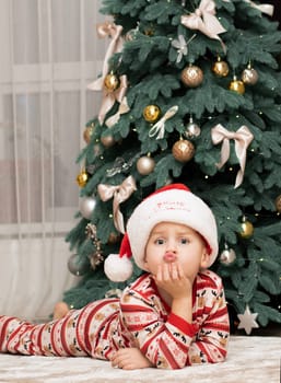 Christmas. A five-year-old boy in New Year's pajamas lies on the floor near the Christmas tree in a Santa Claus hat and smiles cheerfully. Facial expressions of joy and happiness. festive atmosphere