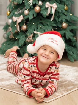 Christmas. A five-year-old boy in New Year's pajamas lies on the floor near the Christmas tree in a Santa Claus hat and smiles cheerfully. Facial expressions of joy and happiness. festive atmosphere
