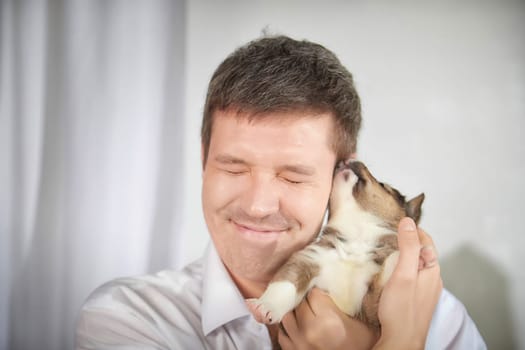 A man is filled with joy as he holds a small puppy close to his face, expressing love and happiness in a well-lit indoor setting.