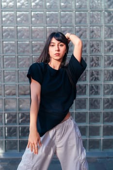 A confident model strikes a dynamic pose in a modern urban environment. The backdrop features glass bricks, enhancing the artistic vibe of the setting. The outfit emphasizes contemporary streetwear.