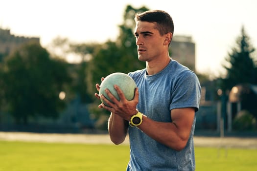 A young male athlete stands on a track field, focused on his upcoming shot put throw. He holds a heavy shot put ball close to his chest while wearing a gray athletic shirt and wristwatch.