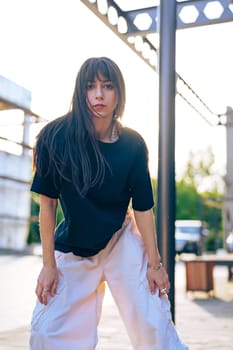 A young woman stands outdoors in an urban area, showcasing a casual outfit with a relaxed posture. The sun sets in the background, creating a warm ambiance.
