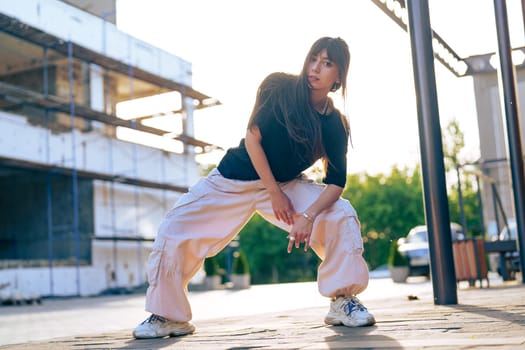 A young woman showcases her dance skills in baggy pants and a black top at a construction site illuminated by the warm glow of sunset. Her confident pose captures the energy of urban dance.