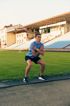 A man in a blue shirt and black shorts is engaged in a squat exercise on a track field. The sun sets, creating a warm glow over the empty stadium seating.
