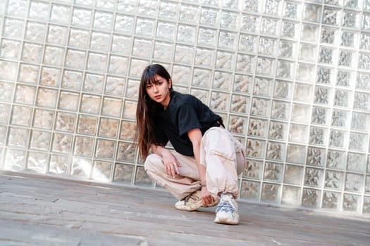 A young woman is crouching in a stylish urban environment, showcasing a dance pose. She wears casual clothing and sports shoes, with sunlight reflecting off a glass brick wall behind her.