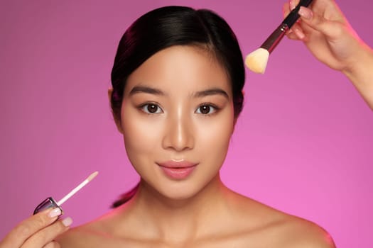 A model is having her makeup applied with glowing skin. One hand holds a brush while another showcases a cosmetic product. The backdrop features a vibrant pink hue.