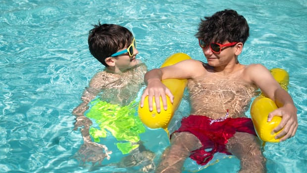 Two boys with sunglasses are enjoying their summer vacation, playing in the pool with an inflatable mattress.