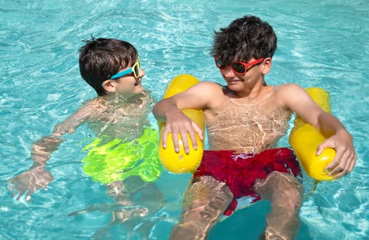 Two boys wearing sunglasses enjoy summer holidays in swimming pool using inflatable buoys