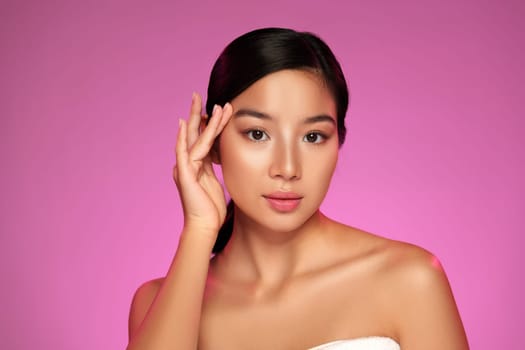 A model exhibits grace with a gentle hand gesture on her face, showcasing natural beauty. The vibrant pink backdrop enhances her features and creates a striking visual contrast.