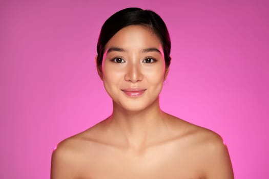 A young woman stands smiling against a bold pink backdrop, radiating confidence and charm. Her fresh makeup and gentle expression highlight her natural beauty in this warm setting.
