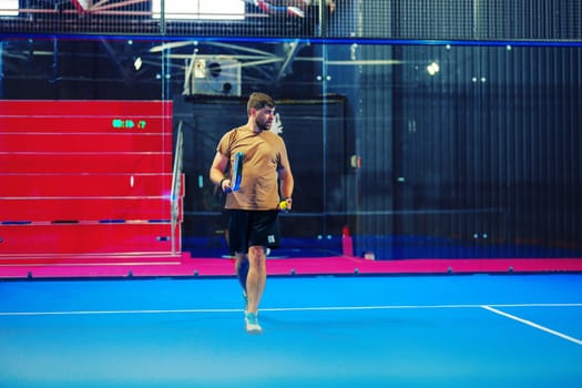 A man is focused on practicing badminton in a well-lit indoor sports center. The vibrant blue court contrasts with red seating, creating an energetic atmosphere for training.