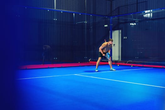 A player stands on a vibrant blue court, preparing to serve in a game of paddle tennis. This indoor facility features glass walls, enhancing the sporty atmosphere.
