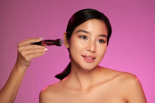 A model is having her makeup applied using a brush in a studio with a soft pink backdrop. The makeup artist focuses on enhancing her natural features.