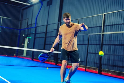 A man is focused on practicing his tennis skills on a vibrant blue indoor court. He prepares to swing his racket as a yellow ball approaches, aiming for precision.
