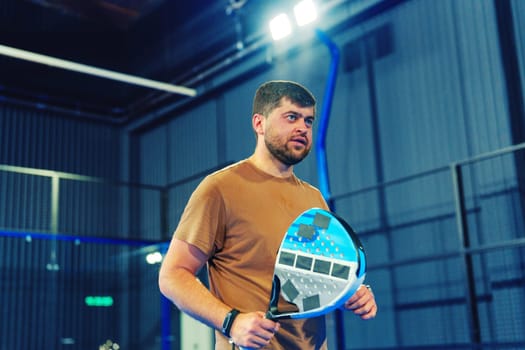 A man stands in a contemporary indoor sports facility, holding a paddle as he prepares for a game. The setting features well-lit courts and modern equipment, ideal for training.