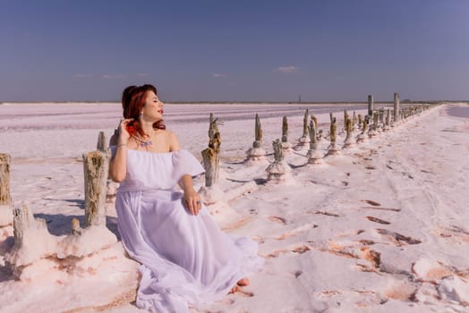A woman in a white dress sits on a wooden fence overlooking a vast salt flat. The sun shines brightly, casting a warm glow on the scene.