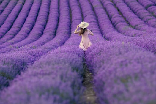 A woman is walking through a field of purple flowers. She is wearing a white hat and a pink dress