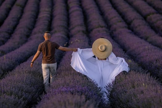 Here's a caption describing the image in under 150 characters: Man and woman stroll hand-in-hand through a vast, vibrant lavender field. She wears a wide-brimmed hat and flowing white fabric.