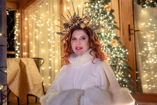 A woman with red hair and a crown of twigs stands in front of a decorated Christmas tree and string lights.