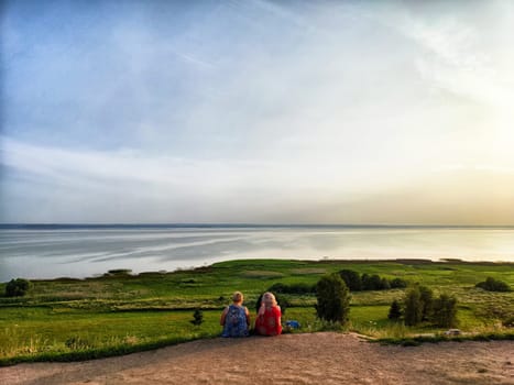 Two people sit side by side on a hillside, admiring the tranquil waters and lush greenery as the sun sets.