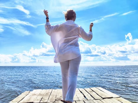 Blonde woman in a white outfit stands on a dock by the water, embracing a carefree moment under the blue sky.