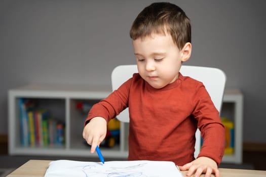 A cute attentive little toddler boy of two years old draws with markers in the album in the children's room at home. Educational activities for kids