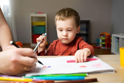 A cute little toddler boy of two years old with dad draws with markers in the album in the children's room at home. Spending time with children. Educational activities for kids