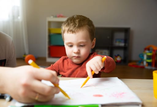 A cute little toddler boy of two years old with dad draws with markers in the album in the children's room at home. Spending time with children. Educational activities for kids