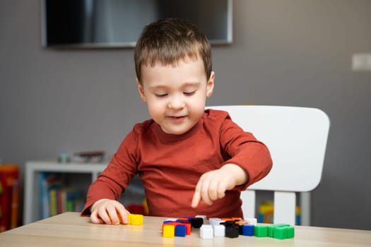 A cute happy little toddler boy of two years old sits at a children's table and plays with multi-colored blocks in his room. Educational toys for children. Soft selective focus