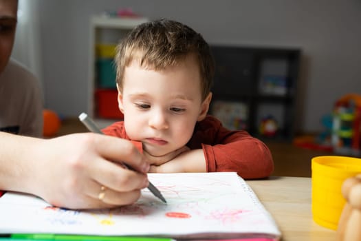 A cute little toddler boy of two years old with dad draws with markers in the album in the children's room at home. Spending time with children. Educational activities for kids