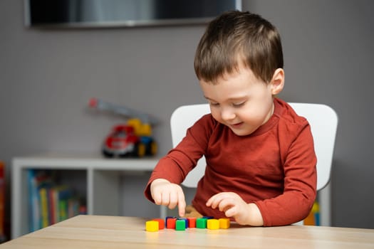A happy cute little toddler boy of two years old sits at a children's table and plays with colorful cubes. Educational logic toys for children. Soft selective focus