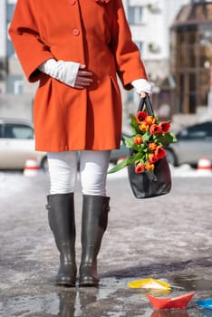Closeup of woman in a orange coat and rubber boots walking down the street with a bouquet of tulips in spring city