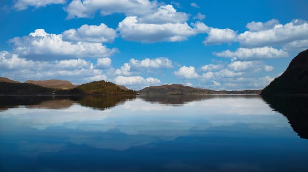 Fjord with view of mountains and fjord landscape in Norway. Blue sky, white clouds, calm mirror-smooth water. Scandinavian landscape shot
