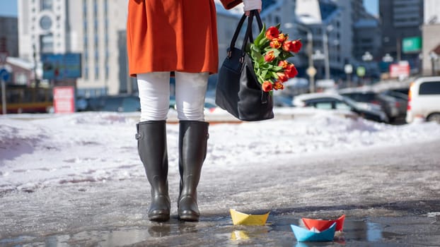 Closeup of woman in a orange coat and rubber boots walking down the street with a bouquet of tulips in spring city
