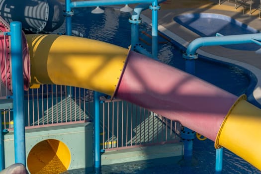 A yellow and pink water slide curves down towards a pool, seen from above. The slide is surrounded by blue and green metal supports and a metal fence.