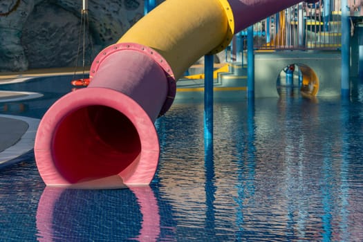 A bright pink and yellow waterslide extends over a blue pool. The end of the slide is submerged in the water, with the opening of the slide visible.