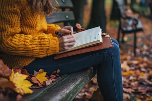 A woman is sitting on a bench and writing in a notebook. The notebook is brown and has a leather cover. The woman is wearing a yellow sweater and jeans. The bench is surrounded by fallen leaves
