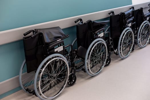 A row of empty folded black wheelchairs along a corridor with a blue wall.