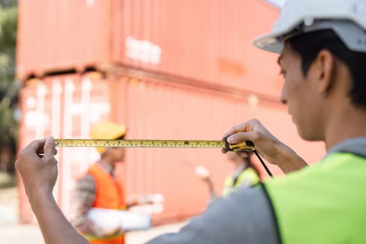 A focused construction worker in a yellow safety vest uses a tape measure to ensure accuracy, while teammates in background discuss blueprints and project details in front of shipping containers.